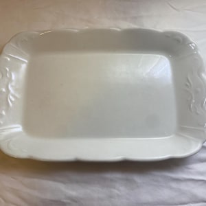 May include: A rectangular, white ceramic serving tray with a scalloped edge and decorative floral accents. The tray has a smooth, glossy surface and is suitable for serving food or displaying items.