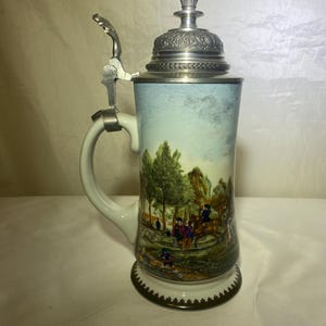 May include: A ceramic beer stein with a hinged metal lid and handle. The stein features a painted landscape scene with figures, trees, and a blue sky. The base has a decorative gold trim.