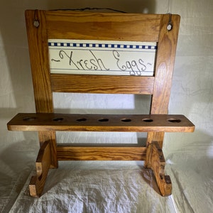 May include: A wooden egg holder with a sign that says "Fresh Eggs" in cursive. The holder has six slots for eggs and is painted a light brown color.