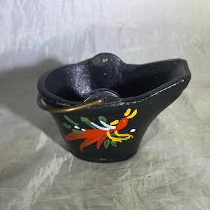 May include: A small black cast iron coal scuttle with a brass handle and painted floral design. The scuttle is decorated with red, yellow, and green flowers.