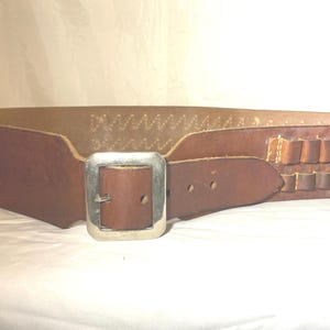 May include: A wide, brown leather belt with a silver-colored buckle. The belt has a series of small, rectangular loops for holding items. The belt is likely for a costume or a Western-themed outfit.
