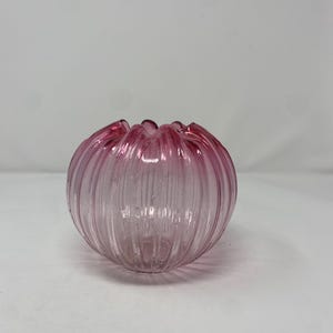May include: A round, pink glass bowl with a ruffled rim. The bowl has vertical ridges and a clear, transparent appearance. The color is a soft, rosy pink, and the bowl is sitting on a white surface.