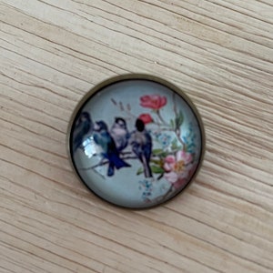 Könnte beinhalten: Ein runder Glascabochon mit einem bronzefarbenen Metallrahmen zeigt eine Vintage-Illustration von drei blauen Vögeln, die auf einem Ast mit rosa und weißen Blüten sitzen.