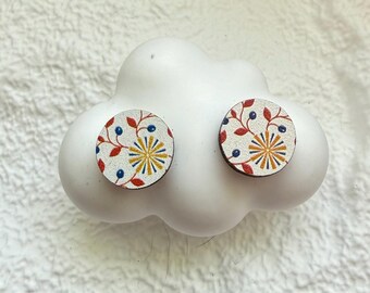 Könnte beinhalten: Ein Paar runde Ohrstecker mit floralem Muster. Die Ohrringe haben einen weißen Hintergrund mit roten Blättern, blauen Beeren und einem gelb-blauen Sternmuster. Sie werden auf einem weißen, wolkenförmigen Objekt präsentiert.