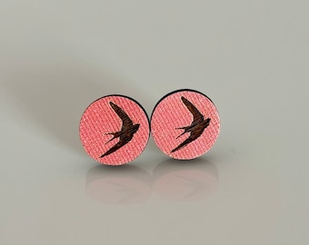 May include: A pair of round stud earrings. Each earring features a dark brown bird silhouette on a pink background. The earrings have a textured appearance and a dark brown border.