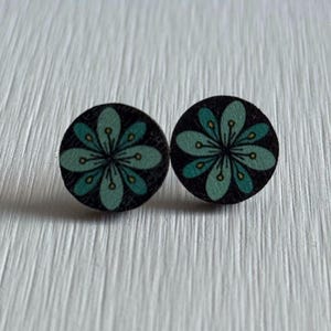 Puede incluir: Un par de pendientes de bot&oacute;n redondos. Cada pendiente presenta un fondo negro con un dise&ntilde;o floral turquesa y amarillo. Los pendientes son de madera y se muestran sobre una superficie blanca texturizada.