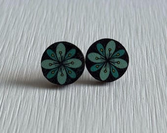 Könnte beinhalten: Ein Paar runde Ohrstecker. Jeder Ohrring hat einen schwarzen Hintergrund mit einem türkis-gelben Blumenmuster. Die Ohrringe sind aus Holz gefertigt und werden auf einer weißen, strukturierten Oberfläche präsentiert.