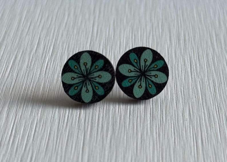Puede incluir: Un par de pendientes de bot&oacute;n redondos. Cada pendiente presenta un fondo negro con un dise&ntilde;o floral turquesa y amarillo. Los pendientes son de madera y se muestran sobre una superficie blanca texturizada.