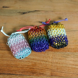 May include: Three small chainmail bags with different color gradients. The bags are made of metal rings and have a drawstring closure. The colors are pink and blue, rainbow, and gold and black.