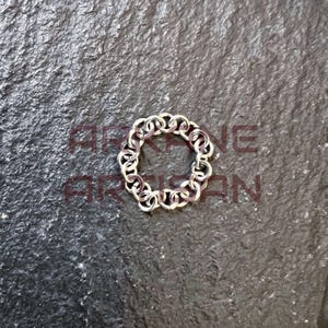 May include: A silver-colored ring composed of interconnected chain links, forming a circular shape. The ring is displayed on a dark, textured surface. The ring's design is intricate and detailed, showcasing a unique style.