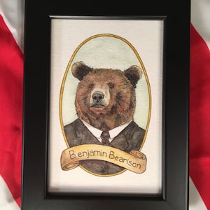 May include: A framed watercolor painting of a brown bear wearing a suit and tie. The bear is in an oval frame with a gold border. The bear's name, "Benjamin Bearison", is written on a scroll below the frame.