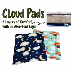 May include: Two square fabric pads labeled Cloud Pads with 5 Layers of Comfort With an Absorbent Layer. One pad features a dark blue background with white clouds, stars, and yellow moons. The other has a light blue background with rainbows and clouds.