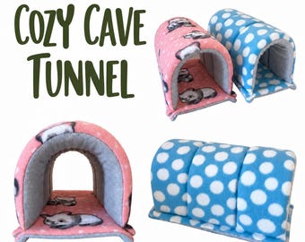May include: Cozy Cave Tunnel text appears next to two soft fleece pet tunnels. One tunnel is pink with a panda print and gray interior, while the other is blue with white polka dots and a gray interior, featuring a curved arch design.