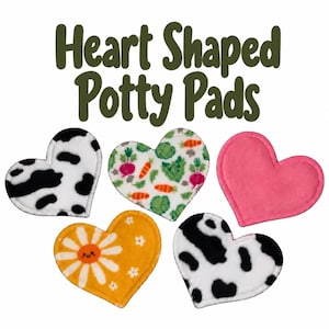 May include: Five heart-shaped potty pads with the text Heart Shaped Potty Pads above them. The pads feature a black and white cow print, a white background with carrots and radishes, a solid pink, and a yellow with a smiling daisy flower.