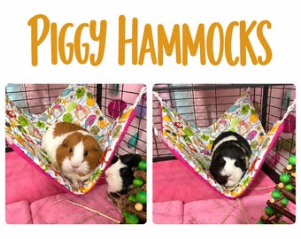 May include: A fabric hammock for small pets with a white background featuring a colorful pattern of guinea pigs, fruits, and leaves. The hammock has bright pink trim and is suspended by white ropes inside a wire cage.
