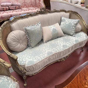 May include: A vintage-style sofa with a gold-colored frame and woven sides. The sofa features a light blue and white patterned seat and back, with matching throw pillows. A round, beige pillow is on the left side.