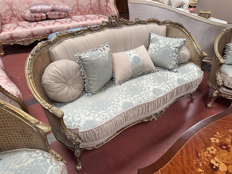 May include: A vintage-style sofa with a gold-colored frame and woven sides. The sofa features a light blue and white patterned seat and back, with matching throw pillows. A round, beige pillow is on the left side.
