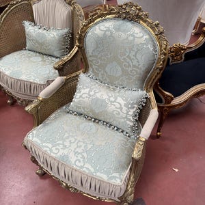 May include: Ornate gold-framed armchair with light blue patterned upholstery and matching pillow. The chair features intricate carvings and a woven cane back. The design is classic and elegant, suitable for a living room or bedroom.