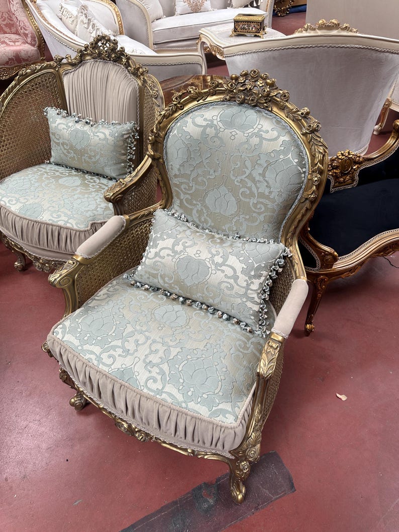 May include: Ornate gold-framed armchair with light blue patterned upholstery and matching pillow. The chair features intricate carvings and a woven cane back. The design is classic and elegant, suitable for a living room or bedroom.