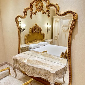 May include: A large, ornate gold-framed mirror dominates the image, reflecting a bedroom scene. The mirror's intricate design complements the gold accents on the bed frame, nightstands, and vanity. A marble-topped vanity and a cushioned bench complete the luxurious bedroom set.