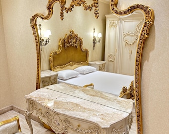 May include: A large, ornate gold-framed mirror dominates the image, reflecting a bedroom scene. The mirror's intricate design complements the gold accents on the bed frame, nightstands, and vanity. A marble-topped vanity and a cushioned bench complete the luxurious bedroom set.