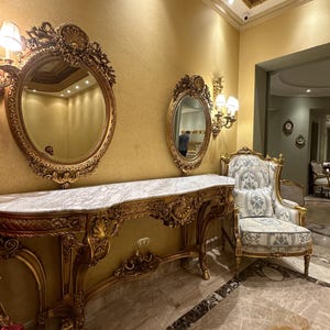 May include: A gold-colored, ornate console table with a marble top, flanked by two oval mirrors with gold frames. A patterned armchair sits nearby. The room has gold-colored walls and a decorative ceiling.