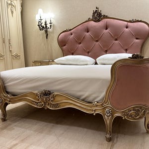 May include: A luxurious bed with a pink, button-tufted headboard and matching side panels. The bed frame is ornate, with gold-colored carvings and curved lines. Two white pillows rest on the bed, which is covered with a light-colored sheet.