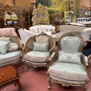 May include: An assortment of antique furniture, including ornate gold-framed chairs with light blue patterned cushions and pillows. Other items include a wooden side table, mirrors, and sofas, all in a vintage style. The background features a large landscape painting.