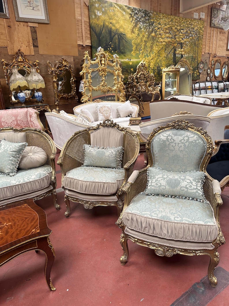May include: An assortment of antique furniture, including ornate gold-framed chairs with light blue patterned cushions and pillows. Other items include a wooden side table, mirrors, and sofas, all in a vintage style. The background features a large landscape painting.