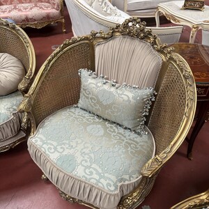 May include: An ornate, antique armchair with a gold-colored frame and woven back. The seat and backrest are upholstered in a light blue floral fabric, complemented by a matching pillow. The chair has decorative carvings and a pleated skirt.