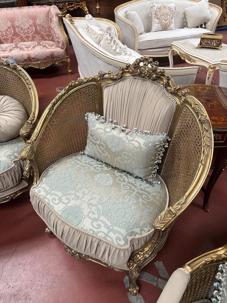 May include: An ornate, antique armchair with a gold-colored frame and woven back. The seat and backrest are upholstered in a light blue floral fabric, complemented by a matching pillow. The chair has decorative carvings and a pleated skirt.