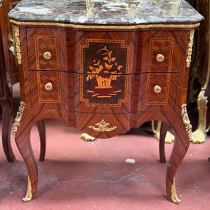 May include: A small, antique wooden cabinet with a marble top. The cabinet features two drawers with inlaid floral designs and gold-colored hardware. The legs are curved and have gold accents, showcasing a classic, ornate style.