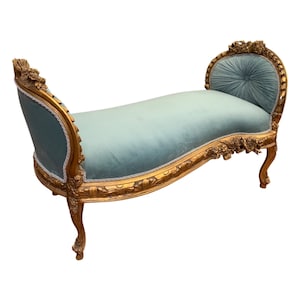 May include: A curved, gold-painted wooden bench with a light blue velvet cushion. The bench has ornate carvings and a tufted back.