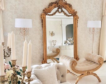 May include: An elegant living room with a large gold-framed mirror, a tufted cream-colored sofa, and a matching chaise lounge. The room features ornate gold sconces, a candelabra, and a floral patterned wallpaper, creating a luxurious atmosphere.