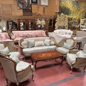 May include: A collection of antique furniture, including sofas, armchairs, and a coffee table. The pieces feature ornate gold frames, light blue and beige upholstery, and decorative details. The setting is a showroom or warehouse.