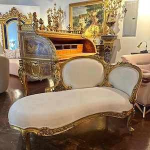 May include: A white chaise lounge with gold-painted ornate details and carved legs. The chaise lounge is upholstered in a white fabric and has a curved back.