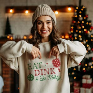 May include: A white sweatshirt with the text "Eat Dink & Be Merry" in green and red. A red paddleball with a green ball is shown next to the text.