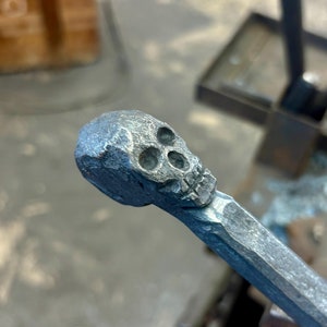 Może przedstawiać: Metalowa czaszka jest kuty na końcu metalowego pręta. Pręt jest zaciśnięty w metalowym imadle.