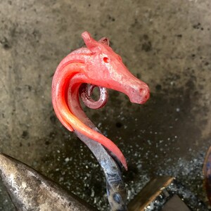 May include: A red-hot metal horse head sculpture is being forged on an anvil. The horse head is being shaped with a hammer and is still hot from the forge.