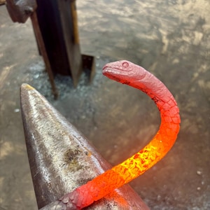 Puede incluir: Una escultura de serpiente de metal al rojo vivo en proceso de forja. La cabeza de la serpiente está detallada con escamas y cuencas oculares. El cuerpo pasa de gris a naranja fuego, indicando el calor del metal.