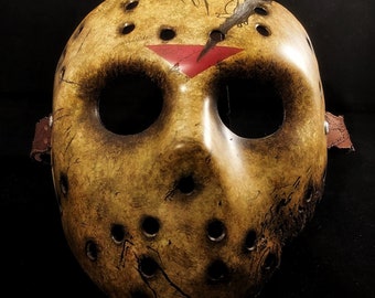 May include: A weathered, yellow hockey mask with a red triangle and black markings on the forehead. The mask has numerous holes and a distressed look.
