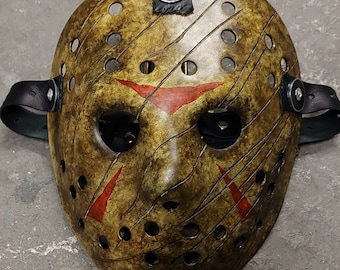 Op de afbeelding: Een bruin en geel hockeymasker met rode verfspatten en zwarte banden. Het masker heeft een verweerde uitstraling en is gemaakt van plastic.