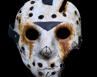 May include: A white hockey mask with black straps and a black triangular design on the forehead. The mask has a distressed look with brown and orange paint splatters and cracks.