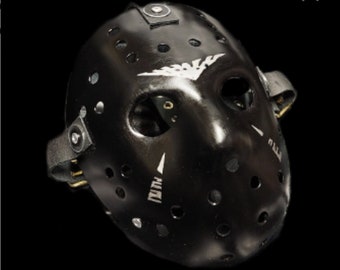 May include: Black hockey mask with a white bird design and multiple holes. The mask has a strap and buckles on the side.