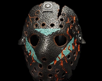 May include: A black hockey mask with a cracked, textured surface. The mask has a teal and orange paint design with a jagged, triangular pattern. The mask has a small black circle on the top.