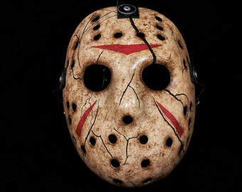 May include: A white hockey mask with red stripes and a cracked, weathered appearance. The mask has a black strap and a small black button on the top.