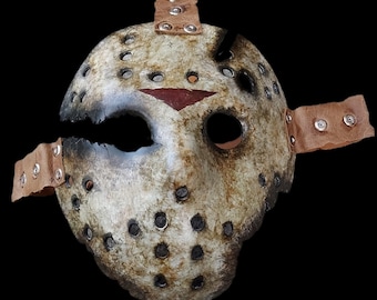 Op de afbeelding: Een wit hockeymasker met een rode streep en bruine leren banden. Het masker heeft veel gaten en een gebarsten uiterlijk.