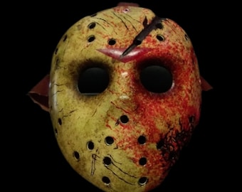 Op de afbeelding: Een wit hockeymasker met rode en gele verfspatten en een zwarte streep over het voorhoofd. Het masker heeft een reeks kleine gaten rond de ogen en de mond.
