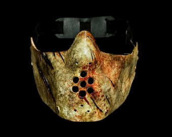 May include: A weathered, distressed mask with a worn, off-white and brown finish. The mask features multiple small circular holes and simulated bloodstains. A black strap is attached for securing the mask.