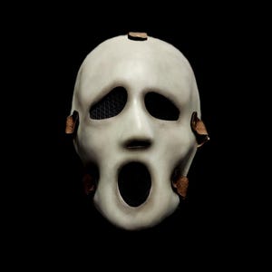 Puede incluir: Una máscara blanca de Scream con aberturas negras para los ojos y la boca. La máscara tiene una apariencia desgastada con parches marrones de cuero adjuntos. La máscara está sobre un fondo negro, creando un contraste dramático.